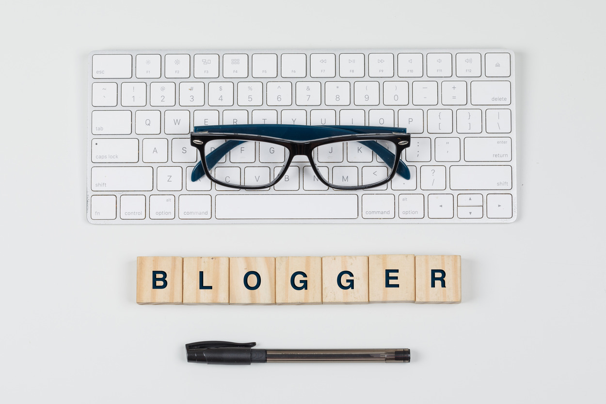 A flat lay of a white keyboard, reading glasses, wooden letter blocks spelling BLOGGER, and a pen on a white background, representing how to optimize blog content for SEO.